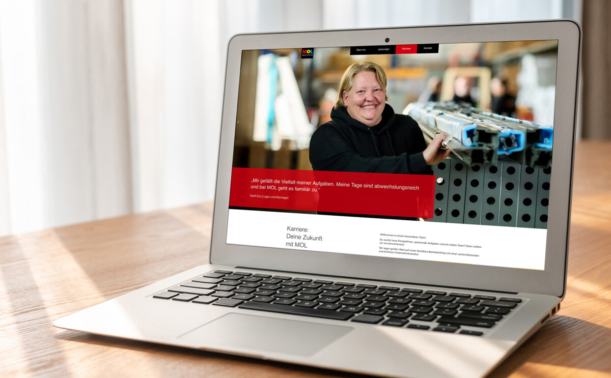 Laptop auf Holztisch zeigt Webseite mit Person in schwarzem Pullover, die Metallteile hält, und rotem Textfeld mit Zitat in deutscher Sprache.
