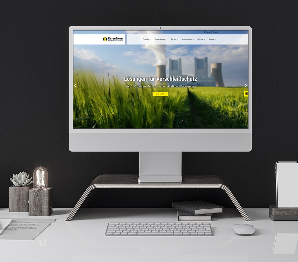 Computerbildschirm zeigt die Webseite von Kalenborn mit dem Text 'Lösungen für Verschleißschutz' vor einem Hintergrund aus grünem Feld und Industrieanlagen.